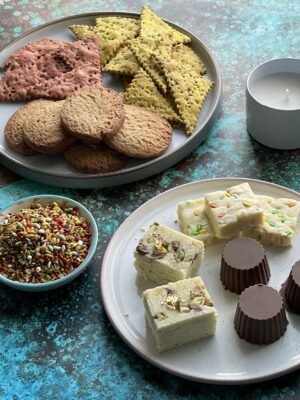 1 candle, a small bowl filled with mukhwas, a larger plate with ginger and cinnamon biscuits and turmeric, jeera crackers alongside some beetroot crackers. A small plate filled with sweets